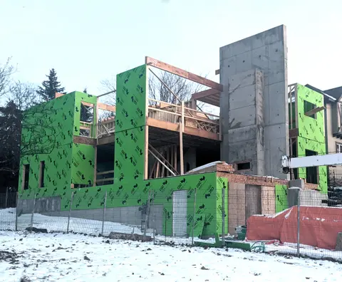 The house under construction in winter, with green sheathing and a tall concrete wall rising through the middle